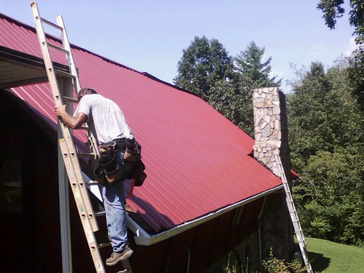 Roofer on a ladder beside a red metal roof and stone chimney — Soffit & Fascia Repair by Roofing Brothers in Muscotah